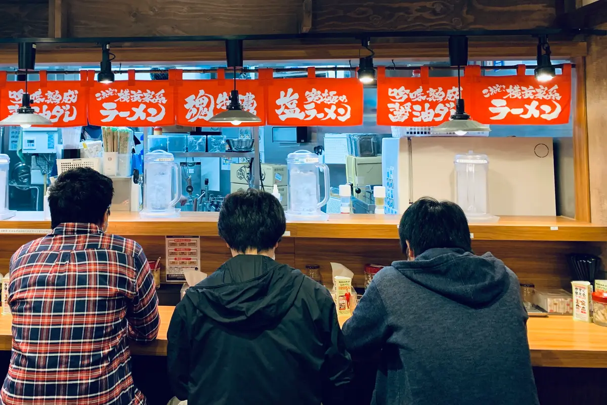 Three customers seen from behind at a ramen counter, facing the open kitchen under a row of red shop curtains