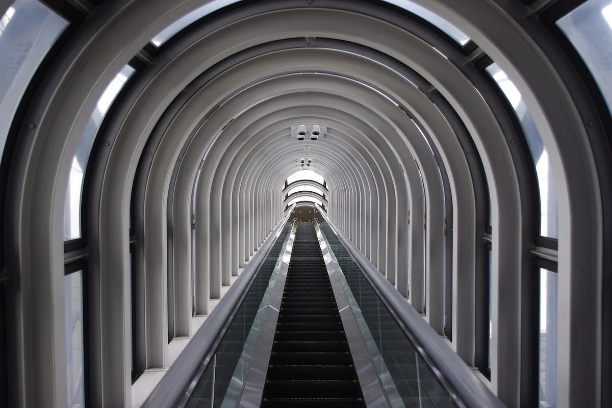 Eine lange Rolltreppe in einer Röhre mit Glaswänden, die durch eine Reihe weißer Bogenrippen zu einer hellen Öffnung oben hinaufführt