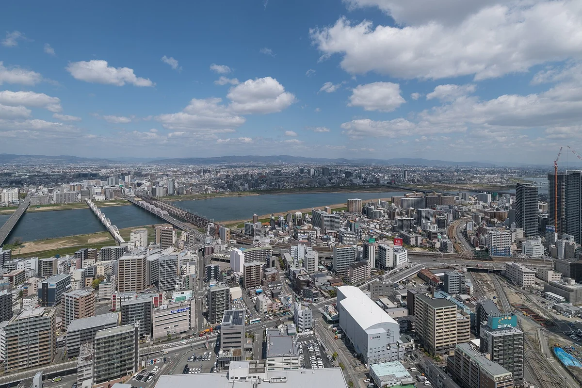 Blick bei Tag von der Aussichtsplattform hoch über der Stadt: Der breite Fluss Yodo mit mehreren Brücken zieht sich durch die Häuser von Osaka, dahinter Berge am fernen Horizont und blauer Himmel mit weißen Wolken