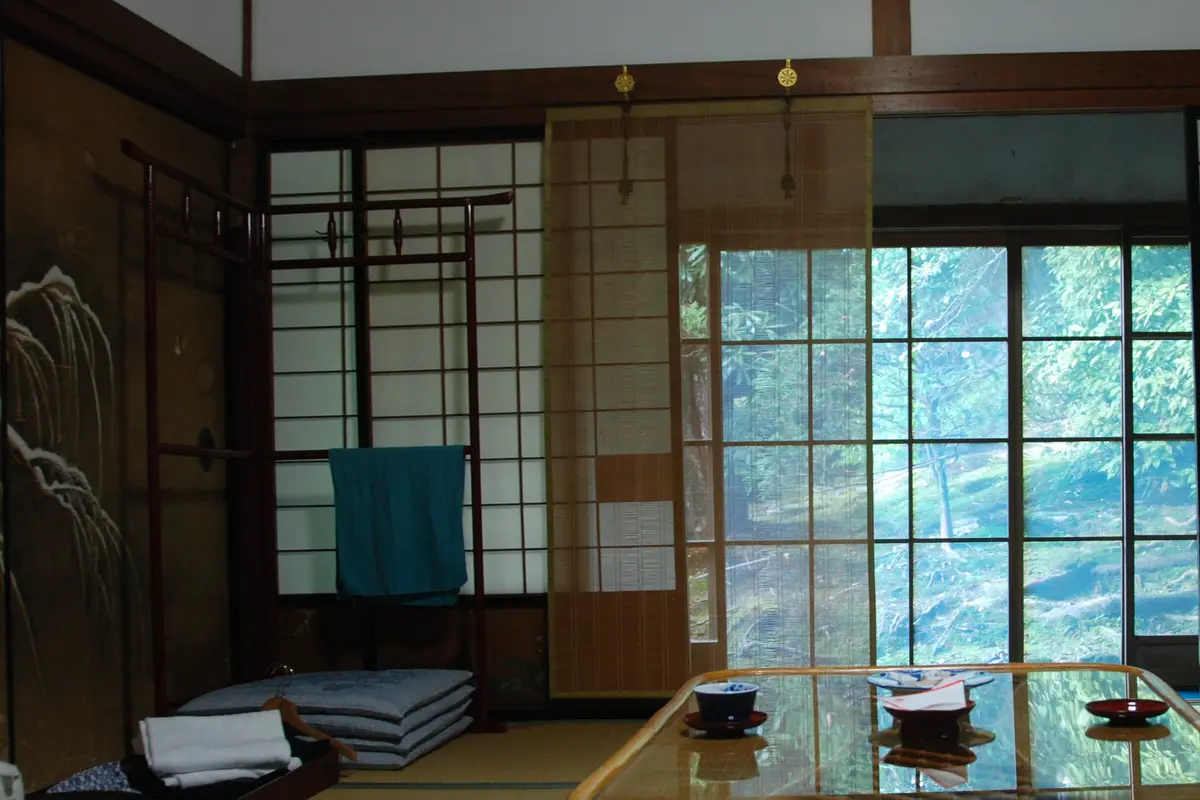 A tatami guest room in a temple lodging, with folded cushions, a tray of towels, tea bowls on a low table and a garden beyond the screens