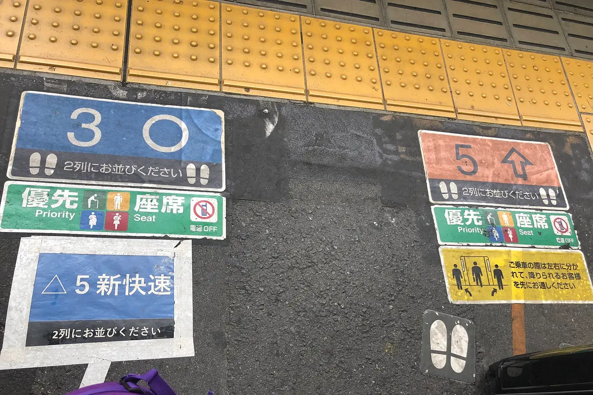 Painted floor markings on a station platform in Osaka, with footprint symbols and Japanese text asking passengers to line up in two rows, beside yellow tactile paving