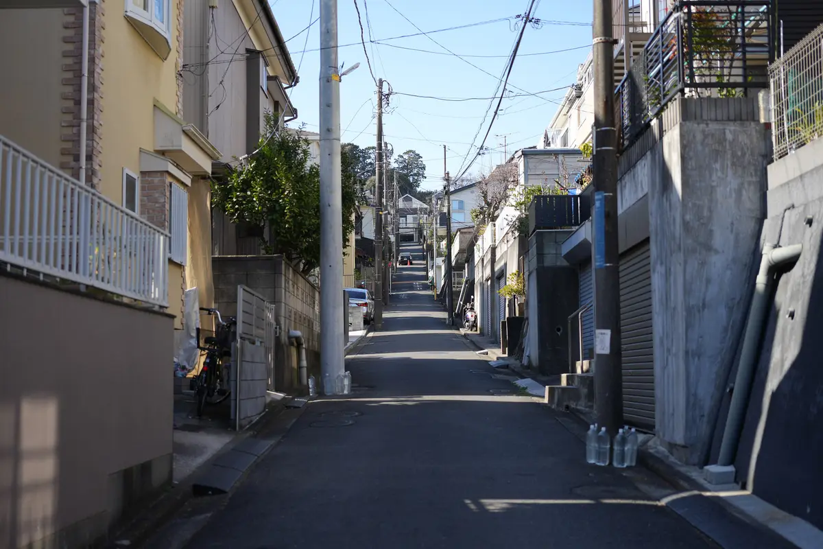 A narrow sloping residential street lined with houses, hedges, a parked bicycle and utility poles, with no traffic on the road