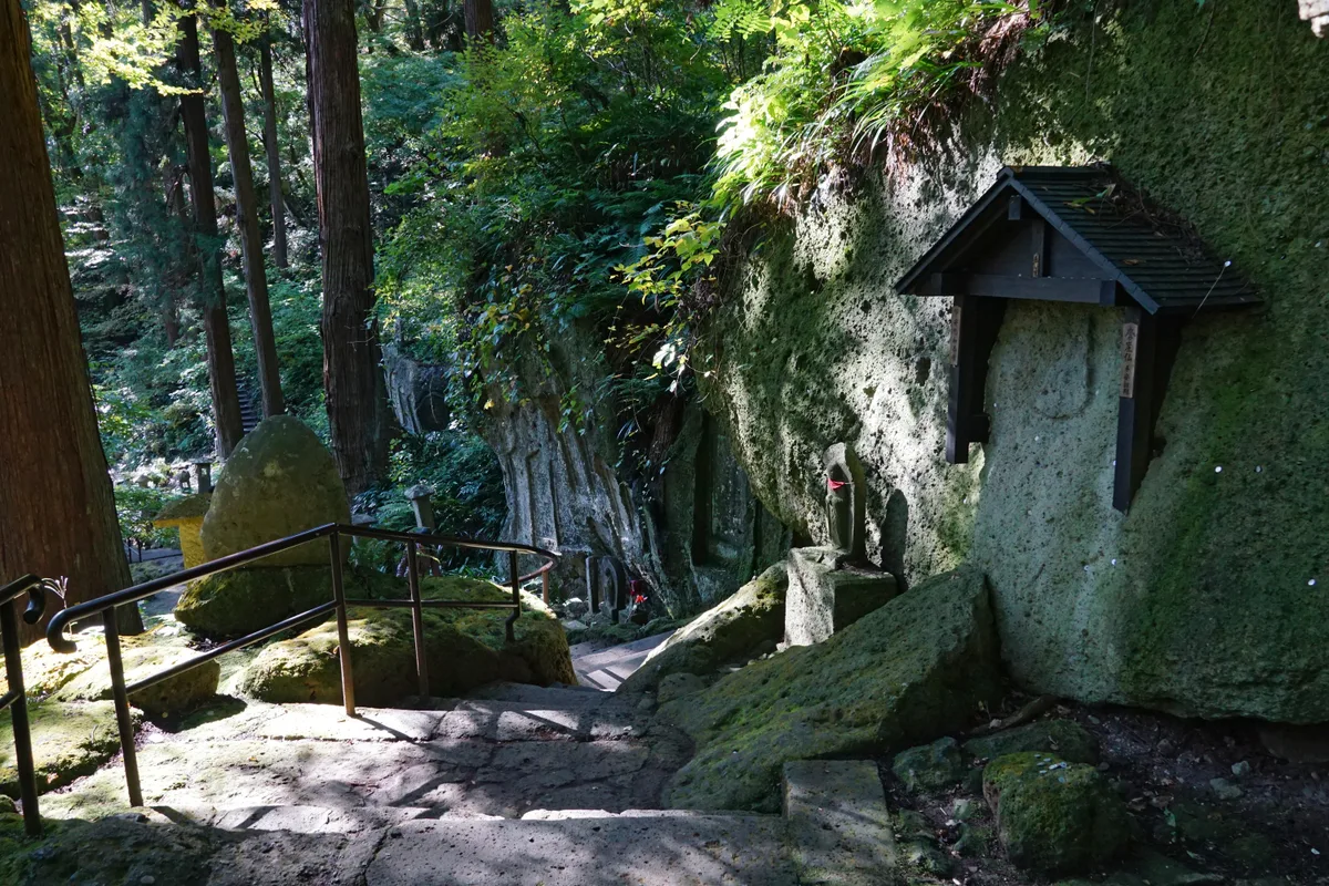Steinstufen mit Metallgeländer neben einer moosbewachsenen Felswand, an der kleine steinerne Jizo-Figuren stehen, eine davon mit rotem Lätzchen, dazu Gravuren im Fels, eine kleine überdachte Holztafel und hohe Zedernstämme