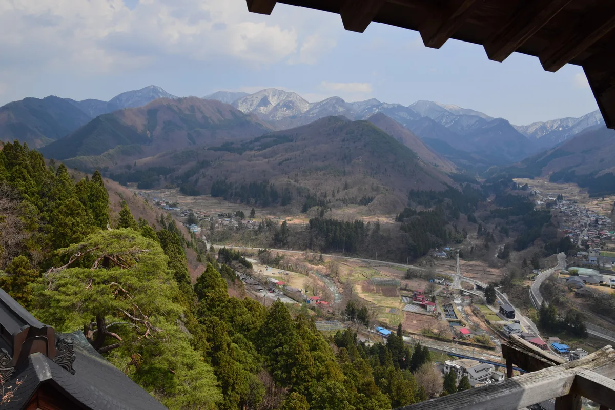 Blick unter dem hölzernen Vordach der Godaido hinaus auf das bewaldete Tal des Tachiya-Flusses, den Ort Yamadera darunter und Berge mit Schnee auf den hinteren Kämmen