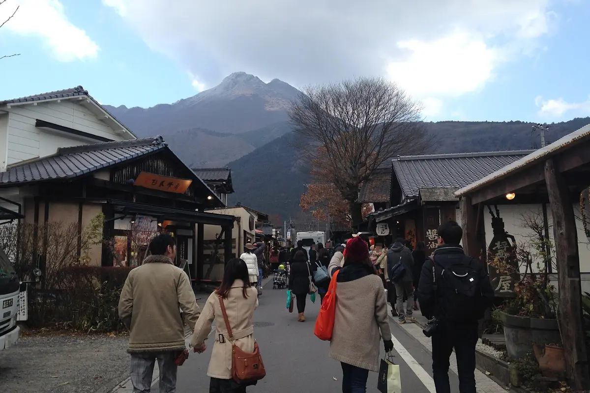 Lo Yunotsubo-kaido visto guardando verso l'alto della via, con persone che camminano tra le facciate in legno dei negozi e, in fondo, le due cime del monte Yufu