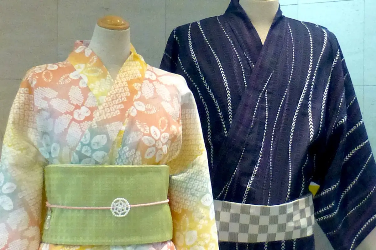 Two headless display mannequins, one in a pastel women's yukata with a green sash, the other in a dark indigo men's yukata with a checkered sash, both with the front panels crossed at the chest