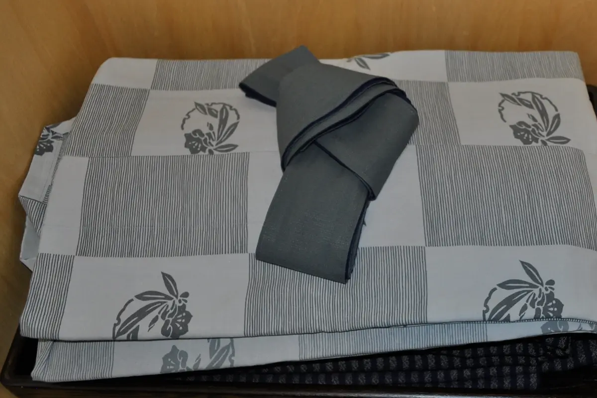 A folded grey and white patterned yukata with a grey sash coiled on top, resting on a dark tray inside a wooden closet