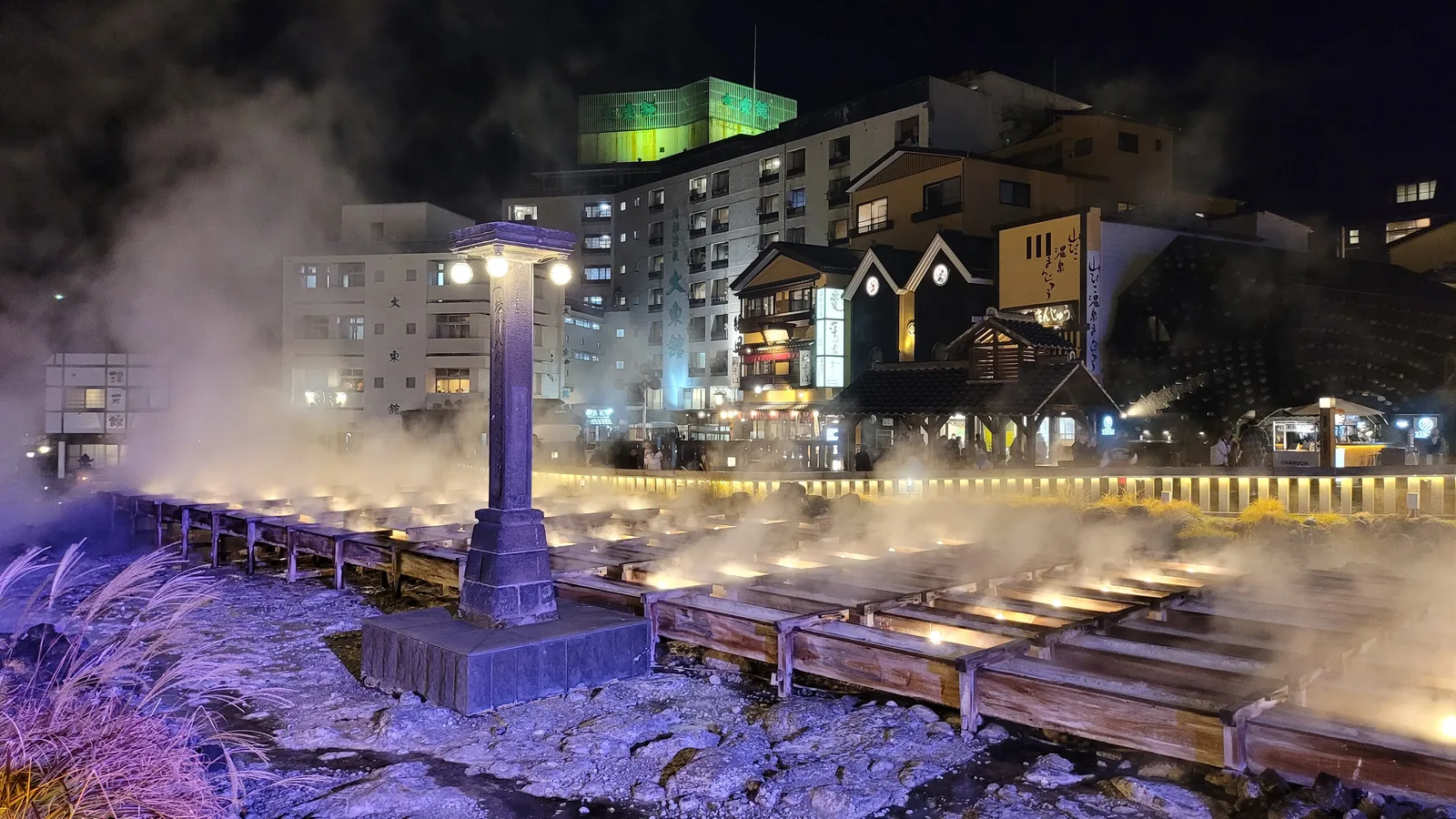 Yubatake at night, its wooden channels lit from below and steam rising around a stone lantern post, with inns lit up beyond