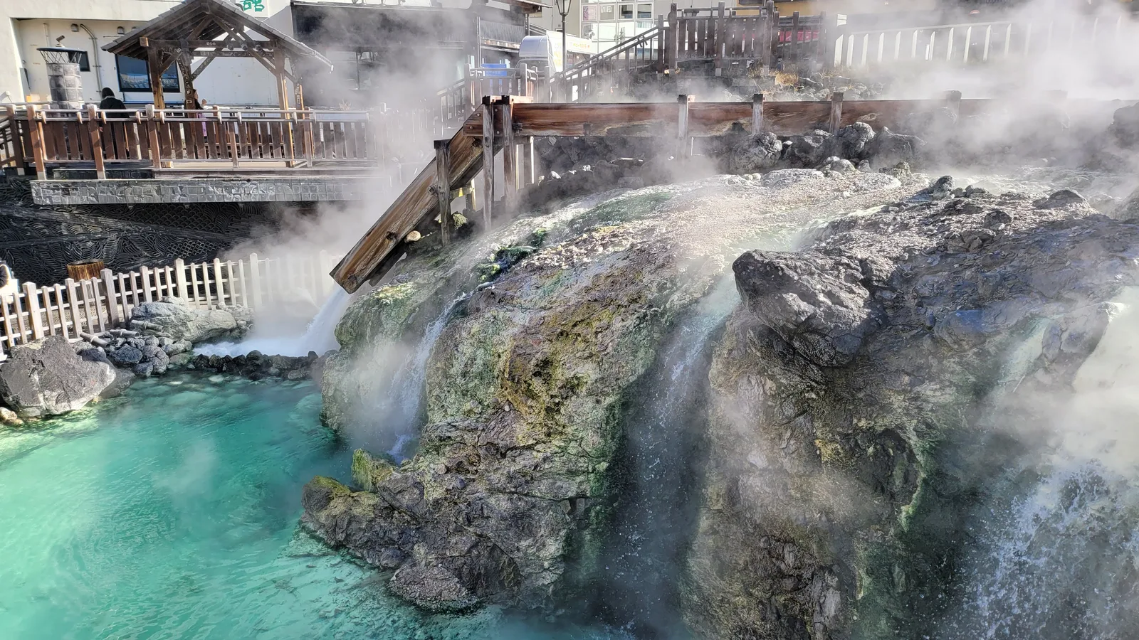 The lower end of Yubatake, where hot spring water from the wooden channels spills over a steaming rock face into a turquoise pool