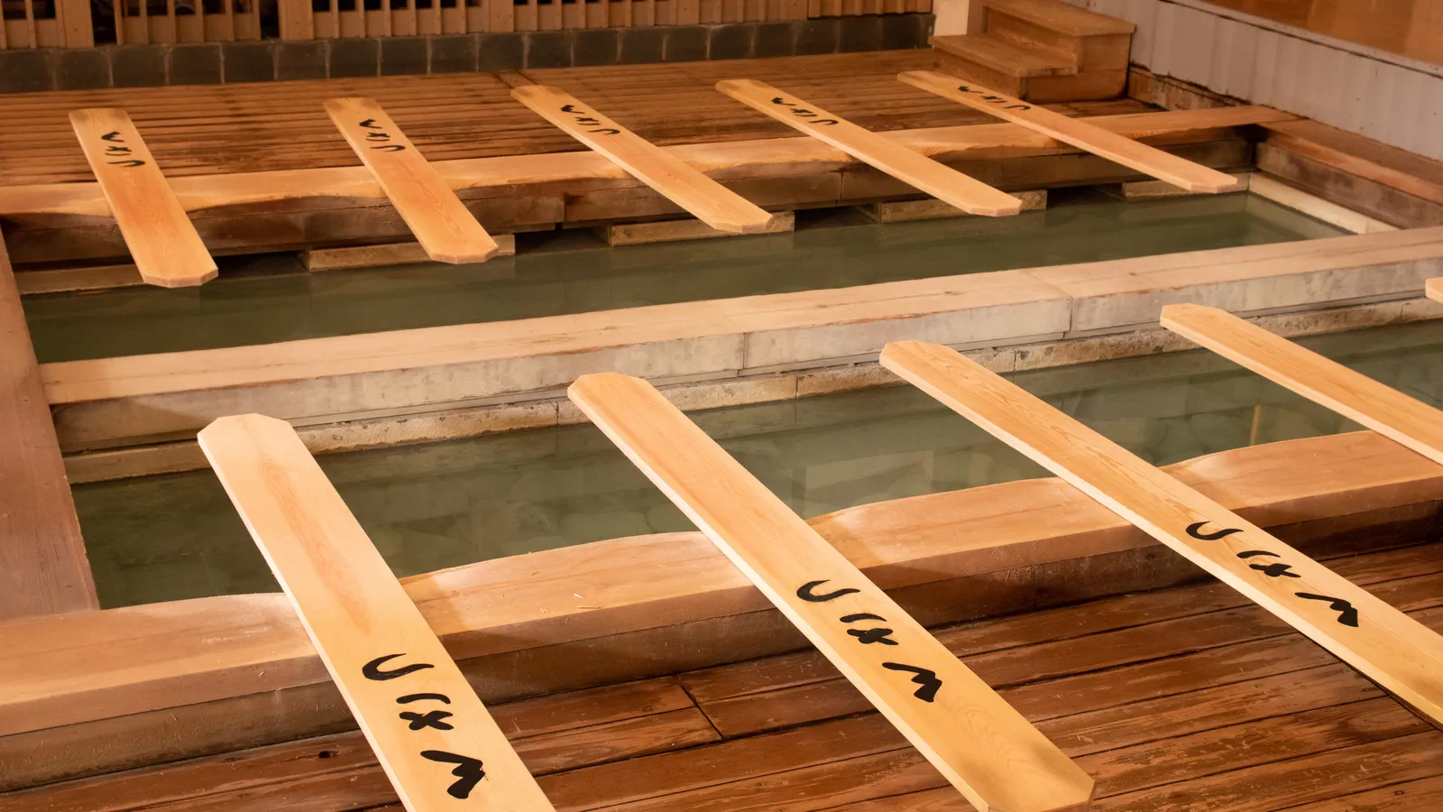 Long wooden yumomi boards with hand-painted lettering, resting across tubs of hot spring water inside the Netsunoyu hall